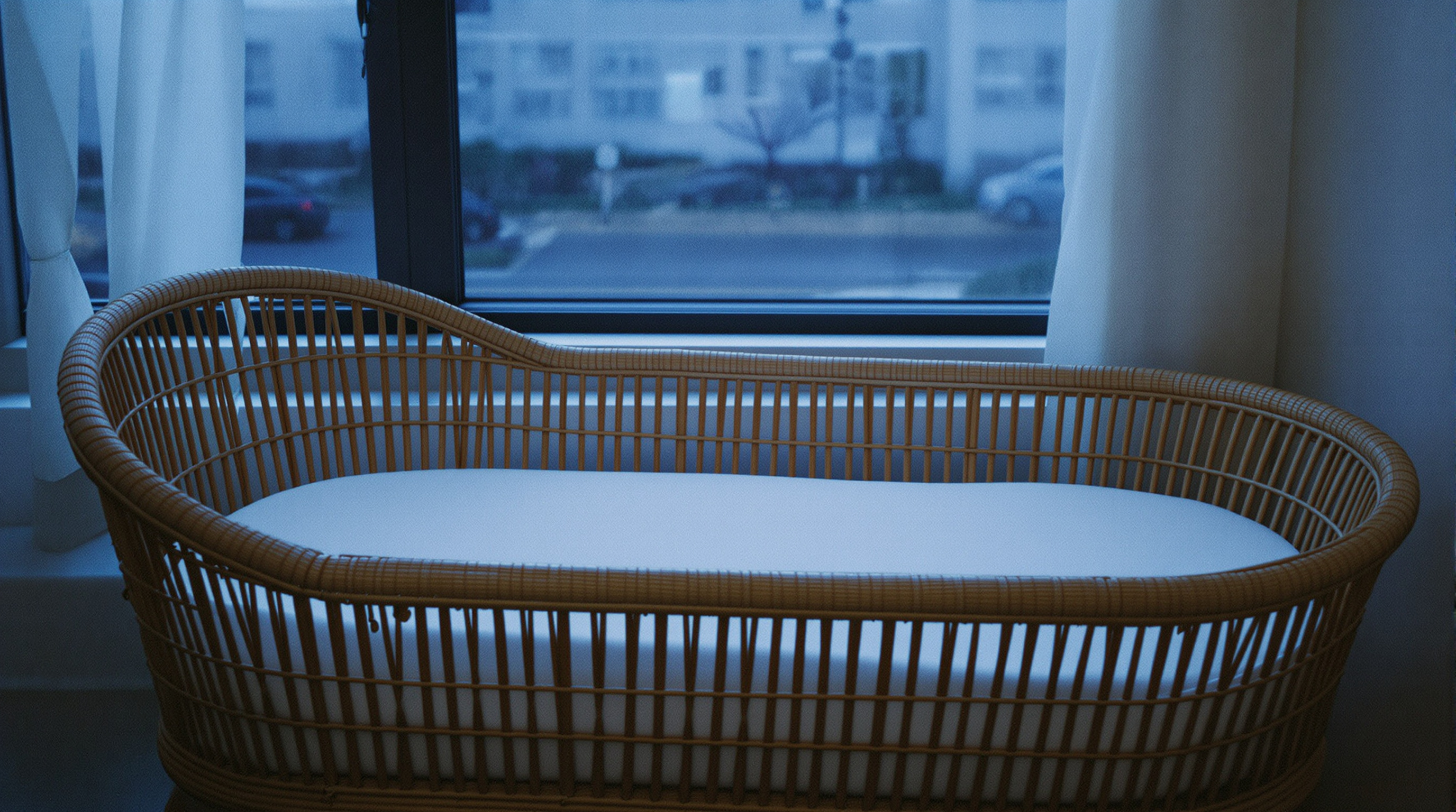 An empty bassinet with a plain fitted sheet beside a window at dusk